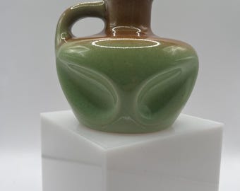 May include: A small, ceramic pitcher with a handle. The pitcher is primarily green with a brown top and handle. The body of the pitcher has a decorative, embossed design. The pitcher is displayed on a white, geometric pedestal.