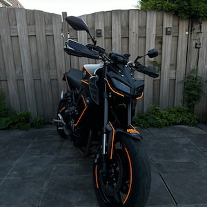 Può includere: Una moto nera con dettagli arancioni. La moto ha una sella nera, un manubrio nero e un parafango anteriore nero. Lo pneumatico anteriore ha una striscia arancione.