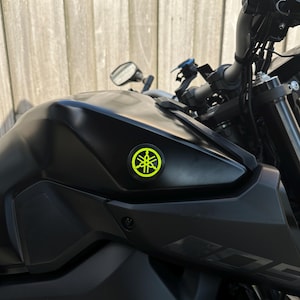 Könnte beinhalten: Ein schwarzes Yamaha-Motorrad mit einem neongrünen Emblem auf dem Kraftstofftank. Das Motorrad hat schwarze Lenker, Spiegel und andere Komponenten. Der Hintergrund ist ein Holzzaun.
