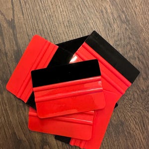 May include: A collection of red and black squeegees, likely for applying vinyl or window tinting. The squeegees have a red, ridged body and a black, rectangular edge. The tools are arranged on a wooden surface.