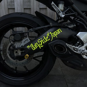Op de afbeelding: Zwarte motorachterwiel met een gele sticker met de tekst "the Dark Side of Japan". De afbeelding toont het wiel, de remschijf en de uitlaat van de motor.