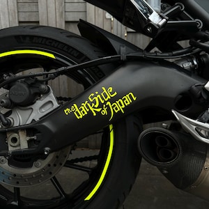 Może przedstawiać: Zbliżenie czarnego tylnego koła motocykla z żółtym paskiem na obręczy. Napis "the Dark Side of Japan" w kolorze żółtym na czarnym wahaczu. Widoczna opona Michelin.