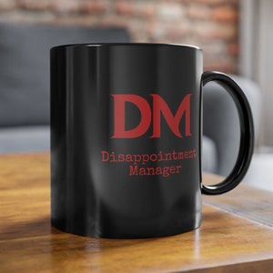 Op de afbeelding: Een zwarte keramische mok met een rood "DM" logo en de tekst "Disappointment Manager".