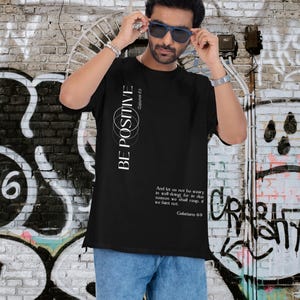 May include: Black t-shirt with the text "BE POSITIVE" and a Bible verse from Galatians 6:9. The shirt has a simple design with white text and is paired with blue denim shorts. The background features graffiti art on a brick wall.