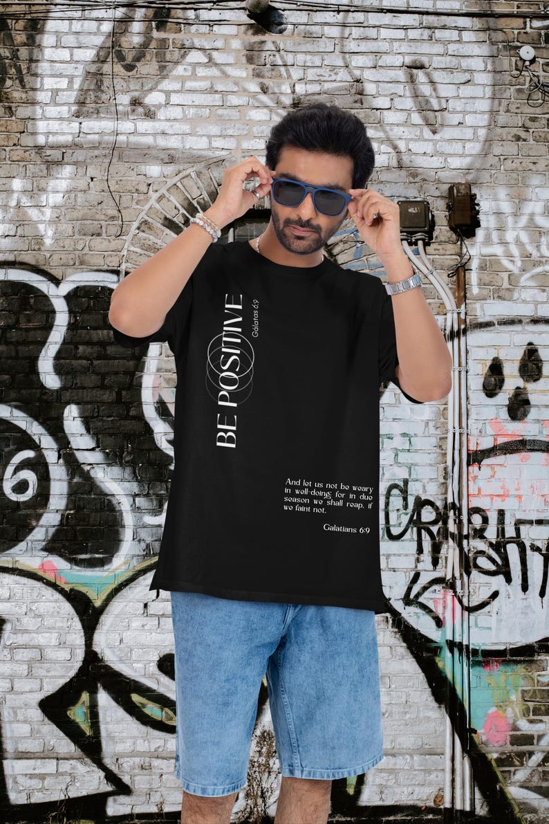May include: Black t-shirt with the text "BE POSITIVE" and a Bible verse from Galatians 6:9. The shirt has a simple design with white text and is paired with blue denim shorts. The background features graffiti art on a brick wall.