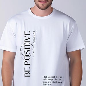 May include: White t-shirt with the words "BE POSITIVE" in black, with a circular design. The text "G&aacute;latas 6:9" is also present. Additional text from Galatians is printed on the shirt. The shirt is being worn by a person.