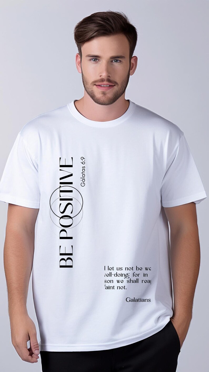 May include: White t-shirt with the words "BE POSITIVE" in black, with a circular design. The text "G&aacute;latas 6:9" is also present. Additional text from Galatians is printed on the shirt. The shirt is being worn by a person.