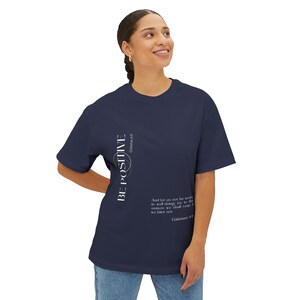 May include: Navy blue t-shirt with the words "BE POSITIVE" in white vertical text. The shirt also features a Bible verse in white text. The t-shirt is a casual, comfortable style.