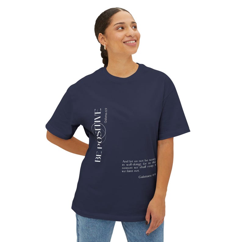 May include: Navy blue t-shirt with the words "BE POSITIVE" in white vertical text. The shirt also features a Bible verse in white text. The t-shirt is a casual, comfortable style.
