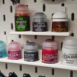 Puede incluir: Una colección de botes de pintura Citadel, organizados en una estantería blanca. Las pinturas son de varios colores, incluyendo naranja, bronce, rojo y azul. Las etiquetas de los botes son claramente visibles, indicando el tipo y color de la pintura.