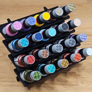 May include: A black paint bottle organiser with multiple tiers, filled with paint bottles. The bottles have colourful lids in shades like pink, blue, yellow, and green. The organiser is made of black material and is placed on a wooden surface.