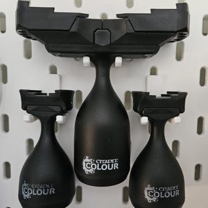 May include: Black Citadel Colour paint bottles and a holder mounted on a white pegboard. The bottles have a teardrop shape and the Citadel Colour logo. The holder is black and designed to hold the bottles.