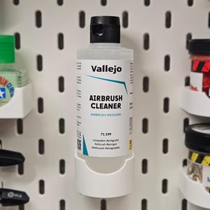 May include: A bottle of Vallejo Airbrush Cleaner, held in a white holder. The bottle has a black cap and a label with the product name and other information. The background is a white pegboard with various art supplies.
