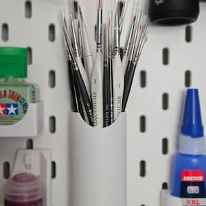 Puede incluir: Una colección de pinceles con mangos blancos y negros, guardados en un soporte cilíndrico blanco. Los pinceles están colocados contra una pared perforada blanca, con varios botes de pintura y pegamento visibles. Los pinceles están etiquetados "RICHMOND".