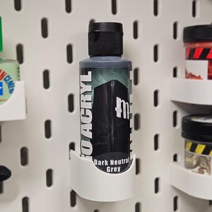 May include: A bottle of "Dark Neutral Grey" acrylic paint is held in a white holder. The paint bottle has a black cap and a label with the brand name and a dark graphic. The holder is mounted on a white pegboard.
