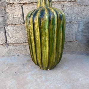 May include: A tall, ribbed vase with a green and black color scheme. The vase has a cylindrical shape with a narrow neck and a wide base. The exterior has vertical ridges, creating a textured surface.