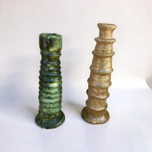 Puede incluir: Dos portavelas cilíndricos con un diseño acanalado y apilado. Uno presenta un esmalte verde oscuro brillante con motas más oscuras, mientras que el otro tiene un acabado texturizado en tonos beige y arena con un brillo sutil.