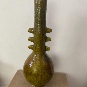Puede incluir: Un jarrón de cerámica artesanal, alto, con base bulbosa y cuello largo y cilíndrico. El jarrón es de color verde oliva moteado con tres juegos de asas redondeadas que sobresalen. El jarrón mide aproximadamente 30 cm de alto.