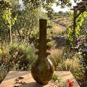 Puede incluir: Un jarrón de cerámica verde oliva, con base bulbosa y cuello cilíndrico texturizado. El jarrón tiene protuberancias decorativas y está sobre una superficie de piedra, con flores silvestres y follaje al fondo. El entorno es exterior.