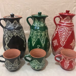 Puede incluir: Un conjunto de tres jarras de cerámica y tres tazas a juego. Las jarras son negras, verdes y rojas, con diseños blancos ornamentados. Las tazas reflejan los colores y patrones de las jarras, mostrando un diseño cohesivo.