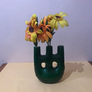 May include: A green ceramic vase with three openings, shaped like a stylized hand, holding a bouquet of yellow and orange flowers.