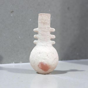 May include: A handmade, off-white ceramic vase with a bulbous base and a narrow neck. The vase features three sets of horizontal protrusions and a small, reddish-pink stain on the base. The vase is likely for decorative purposes.