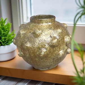 Puede incluir: Un jarrón de cerámica de color dorado, con una superficie texturizada y forma redondeada, con protuberancias redondeadas en relieve. El jarrón tiene un cuello estrecho y está sobre una superficie de madera. Se ve una pequeña planta verde al fondo.