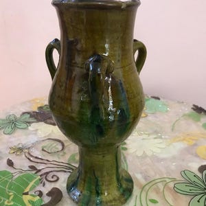 May include: A tall, handcrafted ceramic vase with a dark green and brown glaze. The vase features two loop handles and a flared rim. The base is a pedestal shape. The vase is approximately 12 inches tall.
