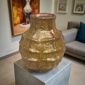 Puede incluir: Un jarrón dorado con textura rugosa, cuerpo ancho y cuello estrecho. Presenta un patrón de formas rectangulares en relieve. El borde interior es de un color cobrizo más oscuro. El jarrón se exhibe sobre un pedestal cuadrado gris. Objeto decorativo.