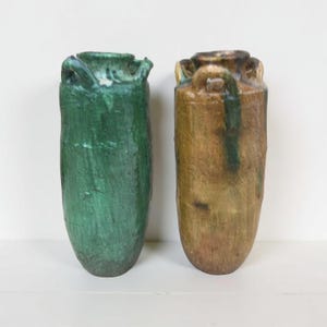 May include: Two ceramic vases. One is a deep green color, the other is a warm brown. Both have a textured surface and a cylindrical shape with a slightly flared top and small handles. The vases are likely for home decor.