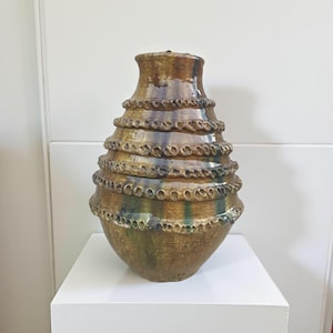 May include: A handcrafted ceramic vase with a unique, tiered design. The vase features a warm, earthy color palette with shades of brown, green, and gold. The textured surface adds visual interest, making it a decorative piece for any home.