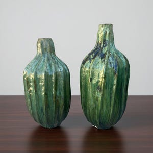 May include: Two green ceramic vases with a textured, ribbed design. The vases have a glossy finish with hints of blue and gold. The vases are of different sizes and have a unique, organic shape, suitable for home decor.