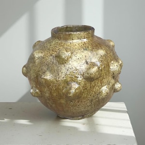 May include: A handcrafted, spherical ceramic vase with a textured, speckled surface and a muted gold color. The vase features evenly spaced, rounded protrusions. The opening is small and circular. The vase is a decorative home accent.