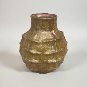 May include: A textured, handcrafted vase with a golden-brown finish. The vase has a bulbous body with a series of raised, square-like patterns and a narrow neck. The interior rim has a reddish-brown hue, adding a subtle contrast to the overall design.