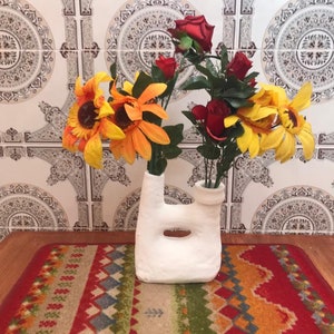 May include: A white ceramic vase with a unique, organic shape holds vibrant sunflowers and red roses. The vase sits on a colorful rug with geometric patterns, against a backdrop of patterned tiles.