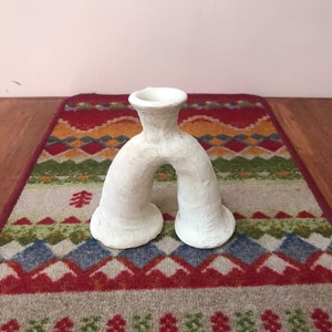 May include: White ceramic vase with a unique, abstract shape. The vase has two openings at the top and a rounded, arching base.