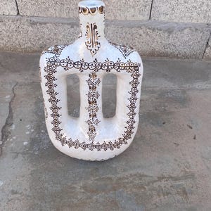 Puede incluir: Jarrón de cerámica blanca con un diseño único de centro abierto. El jarrón está adornado con intrincados patrones marrones y dorados, incluyendo motivos florales y geométricos. Tiene un cuello alto y delgado y una base ancha y redondeada. La estética general es artesanal y decorativa.
