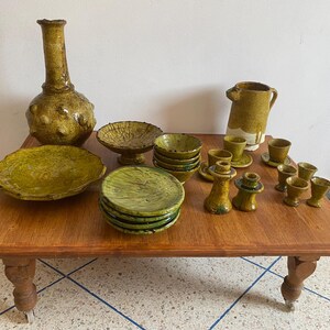 Puede incluir: Una colección de vajilla de cerámica esmaltada en color verde oliva que incluye un jarrón grande texturizado, una jarra, un plato ancho, un cuenco con pie, una pila de platos pequeños, varios cuencos pequeños, dos candelabros y varias tazas pequeñas con platillos.