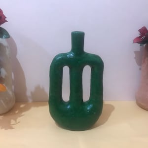 May include: A green ceramic vase with a unique, abstract design. The vase has two cut-out openings on the front and a smooth, glossy finish.