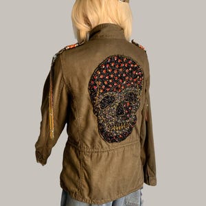 May include: Olive green jacket with a large, beaded skull design on the back. The skull is black with red accents. The jacket has gold-colored chain detailing and epaulettes on the shoulders.