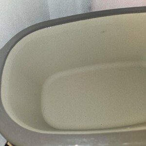 May include: Close-up of a gray ceramic baking dish with a light beige interior. The dish has a rectangular shape with rounded corners and a gray rim. The interior has a smooth, slightly textured surface.