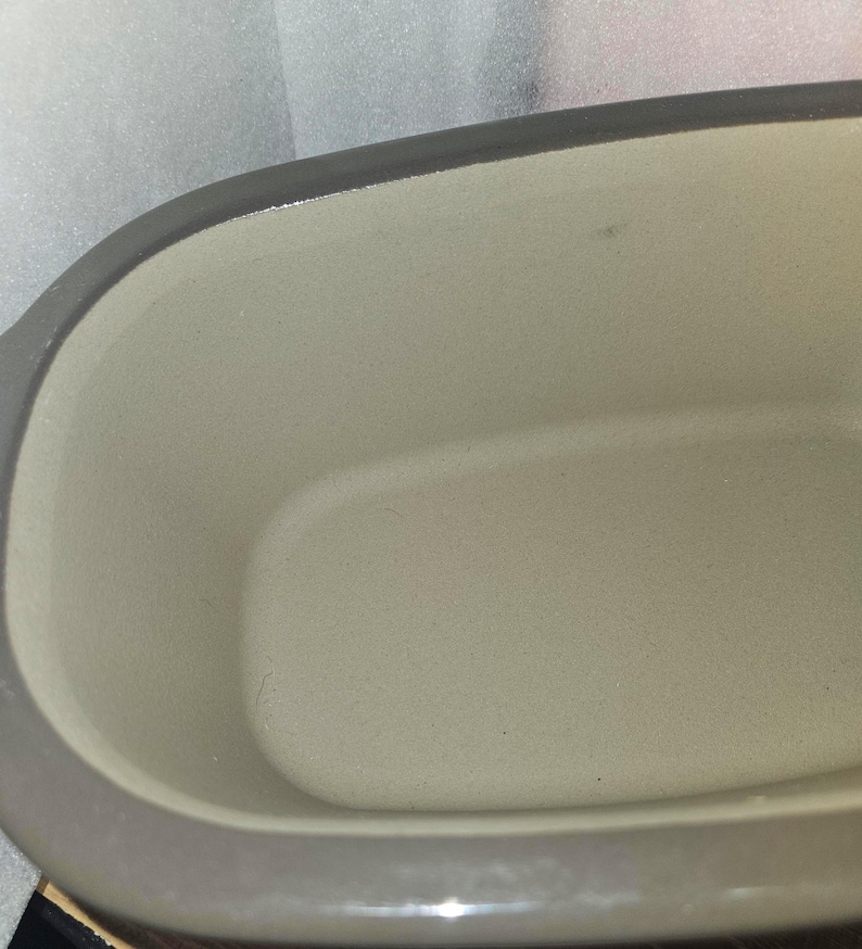 May include: Close-up of a gray ceramic baking dish with a light beige interior. The dish has a rectangular shape with rounded corners and a gray rim. The interior has a smooth, slightly textured surface.