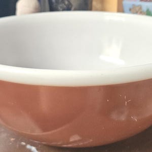 May include: A round, two-tone mixing bowl. The exterior is a warm, reddish-brown color, while the interior is white. The rim of the bowl is also white. The bowl is likely made of ceramic or glass and is designed for food preparation.