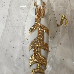 May include: A white porcelain angel ornament with gold accents and a gold ribbon. The angel is wearing a gown with a gold pattern and has wings.