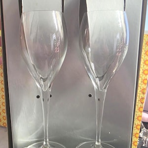 Puede incluir: Un juego de dos copas de champán de cristal transparente con tallos delgados y cuencos redondeados, presentadas en una caja de cartón plateada. La base de cada copa presenta el texto impreso en blanco Laurent-Perrier, Toasting Glasses y Flûtes à champagne.