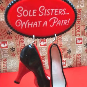 Puede incluir: Adorno navideño rojo y negro con forma de par de zapatos de tacón. El adorno tiene el texto "Sole Sisters... WHAT A PAIR!" en un óvalo rojo. Los zapatos tienen tacones negros y una flor negra.