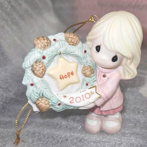 May include: A ceramic Christmas ornament featuring a figurine holding a wreath. The wreath is decorated with pinecones and red berries, and a star with the word "Hope". The year "2010" is also visible. The figurine is wearing a pink coat.