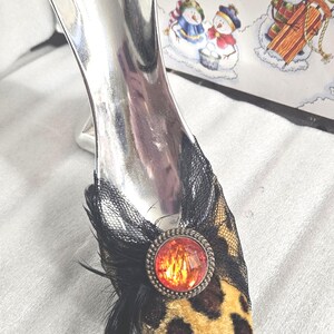 May include: A decorative shoe ornament with a silver heel and a leopard print toe. The toe is adorned with black lace, a red jewel, and black feathers. A winter-themed illustration is in the background.