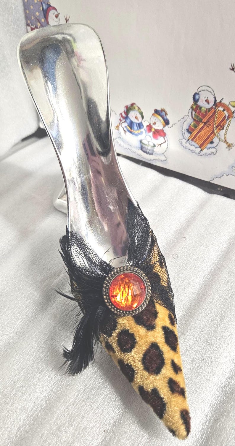 May include: A decorative shoe ornament with a silver heel and a leopard print toe. The toe is adorned with black lace, a red jewel, and black feathers. A winter-themed illustration is in the background.