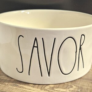 May include: Cream-colored ceramic bowl with the word "SAVOR" in black, handwritten-style lettering. The bowl has a rounded shape and a smooth, glossy finish. The interior is a lighter cream color.